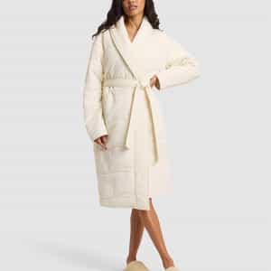 A woman wears the Cozy Earth Women's Quilted House Coat in Vanilla, featuring a waist tie and matching slippers, as she stands facing forward against a plain light gray background. |Color:Vanilla