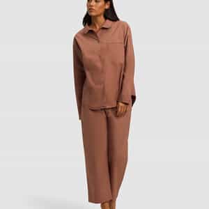 A woman stands barefoot against a plain background, wearing the Cozy Earth Women's Sutton Long Sleeve Pajama Set—a loose-fitting brown button-up top with a chest pocket and matching wide-leg pants.|Color:Saddle