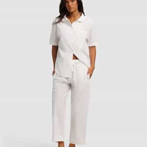 A woman stands barefoot in the Cozy Earth Women's Sutton Short Sleeve & Pant Pajama Set, featuring a matching white button-up shirt and relaxed-fit pants. She has long dark hair, hands in her pockets, against a plain light gray background.|Color:White