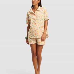 A woman stands barefoot in the Cozy Earth Women's Sutton Short Sleeve Pajama Set, featuring a loose button-up shirt and matching shorts with a pastel floral pattern, looking relaxed against a plain background.|Color:Wildflower