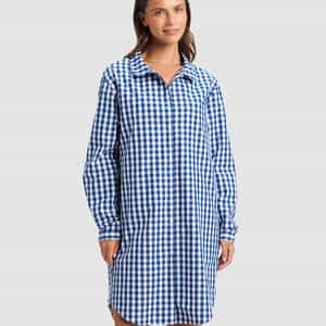 A woman with straight brown hair wears the Cozy Earth Women's Sutton Sleep Dress, a blue and white checkered, long-sleeve shirt dress that falls above the knees. She smiles slightly against a plain light background.|Color:Pacific Gingham