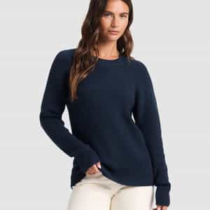 A woman with long brown hair models the Cozy Earth Women's Classic Crewneck in navy blue, paired with off-white pants. She stands against a plain light gray background, one hand in her pocket.|Color:Eclipse