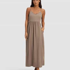 A woman models the Cozy Earth Women's Sunset Gathered Dress, an ankle-length, sleeveless taupe dress with thin straps and a gathered waist. She wears black sandals and poses with one hand in her pocket against a light background.|Color:Ebony