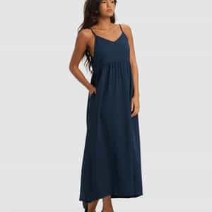 A woman with long dark hair stands against a plain background wearing Cozy Earth's Women's Sunset Gathered Dress - Last Chance, a sleeveless navy blue, ankle-length dress with thin straps and black flats, one hand in her pocket.|Color:Eclipse