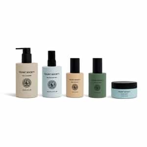 Five skincare products from Cozy Earth’s The Routine are arranged in a row, including a gel cleanser with a pump, a balancing mist spray, two bottles with black caps, and a small blue jar with a black lid.