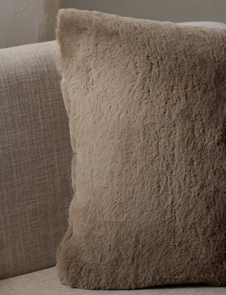 The Cozy Earth Cuddle Pillow, a square beige fuzzy throw, sits on a light fabric sofa, adding cozy softness to your neutral-toned space.|Color:Walnut |Size:24" x 24"