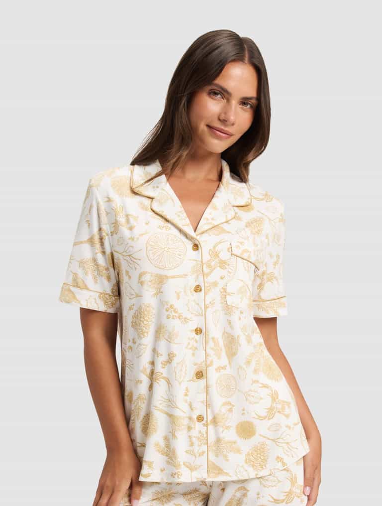 A woman with long brown hair models the Cozy Earth Women's Bamboo Stretch-Knit Short Sleeve Pajama Top, part of a white pajama set with gold botanical prints. She pairs it with matching shorts and smiles softly against a plain light background.|Color:Winter Toile Dijon