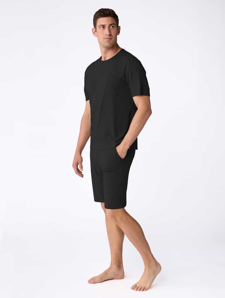 A man stands barefoot on a white background, wearing the Cozy Earth Men's Bamboo Stretch-Knit Short Sleeve Pajama Set in black, featuring a matching short-sleeve T-shirt and pocketed shorts, looking slightly to his left. |Color:Black