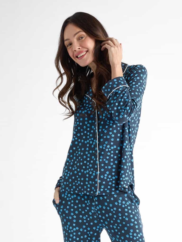 A smiling person in Cozy Earth Women's Bamboo Stretch-Knit Long Sleeve Pajama Top, featuring a dark blue fabric with small blue floral patterns, stands with one hand in their pocket against a white backdrop. |Color:Forget Me Nots