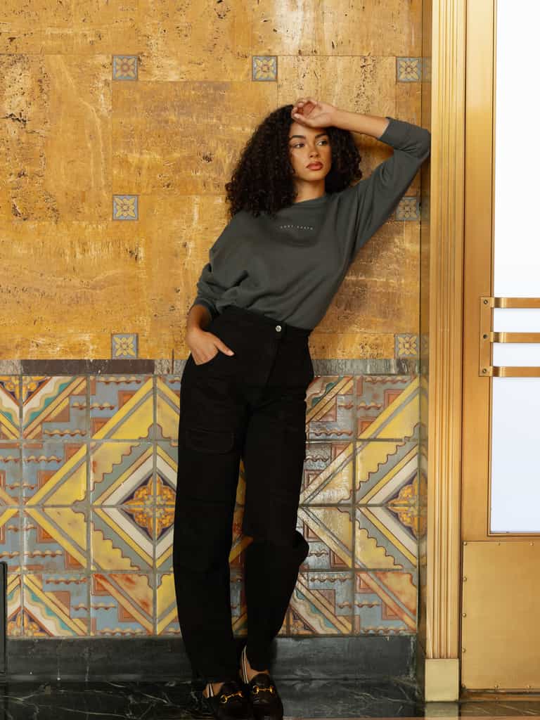 Storm CityScape Pullover Crew. The Pullover is being worn by a female model. An accompanying pair of black jeans are being worn to complete the look of the outfit. The photo was taken with a city building background. |Color:Storm