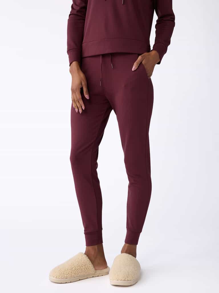 A person is wearing Cozy Earth's Women's Bamboo Jogger Pants in burgundy with their hands in the pockets, paired with beige fuzzy slippers, standing against a plain white background. |Color:Burgundy