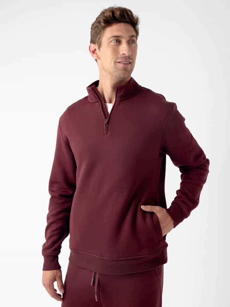 A man stands against a plain background, wearing the Cozy Earth Men's CityScape Quarter Zip in maroon with one hand in his pocket. He has short brown hair and is looking slightly to the side with a relaxed expression. He is also wearing matching maroon sweatpants. |Color:Burgundy