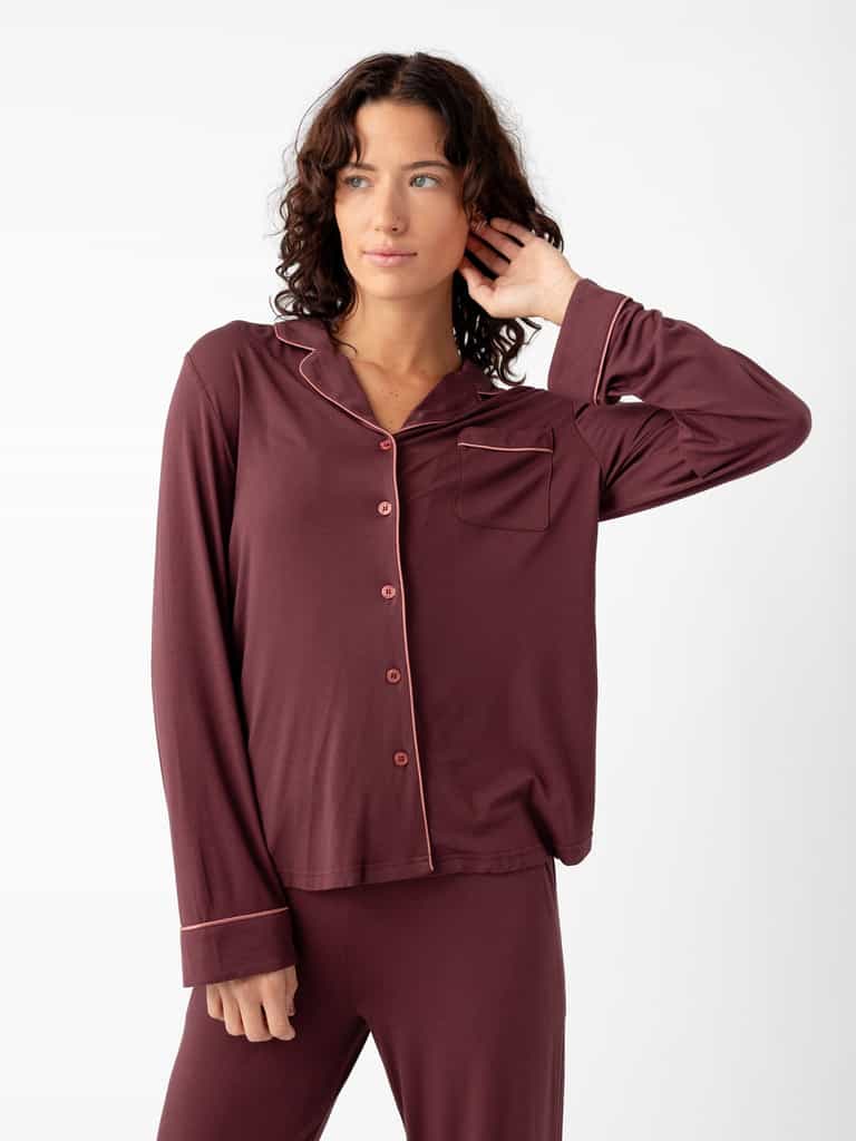 A person in the Cozy Earth Women's Bamboo Stretch-Knit Long Sleeve Pajama Set poses against a plain white background. The burgundy pajamas feature buttons, pocket, and contrast piping. They touch their hair with one hand and look slightly to the side. |Color:Burgundy