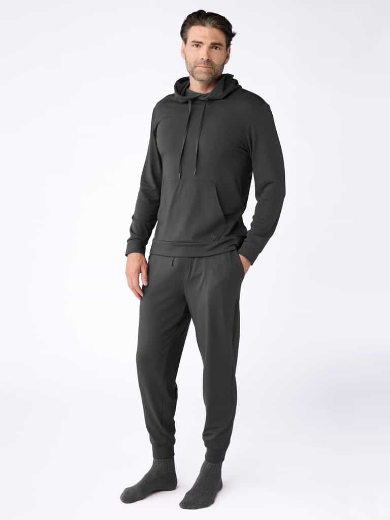 A man wears the Cozy Earth Men's Brushed Bamboo Hoodie & Jogger Set in dark gray, with his hands in his pockets and gray socks, posing against a plain white background. |Color:Charcoal
