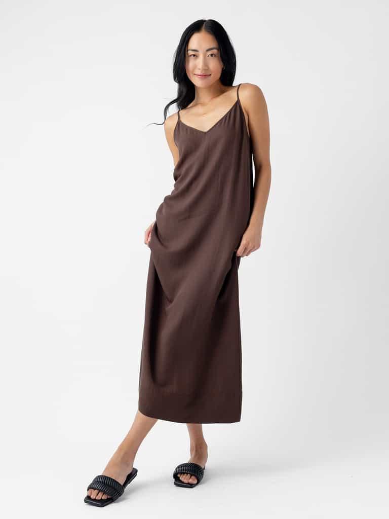 A woman with long, dark hair stands against a white background. She is wearing Cozy Earth's Women's Sunset Slip Dress in a loose, brown spaghetti strap design that reaches her ankles and black slide sandals. She is slightly leaning to one side, with one hand in her pocket and the other touching the dress. |Color:Cacao