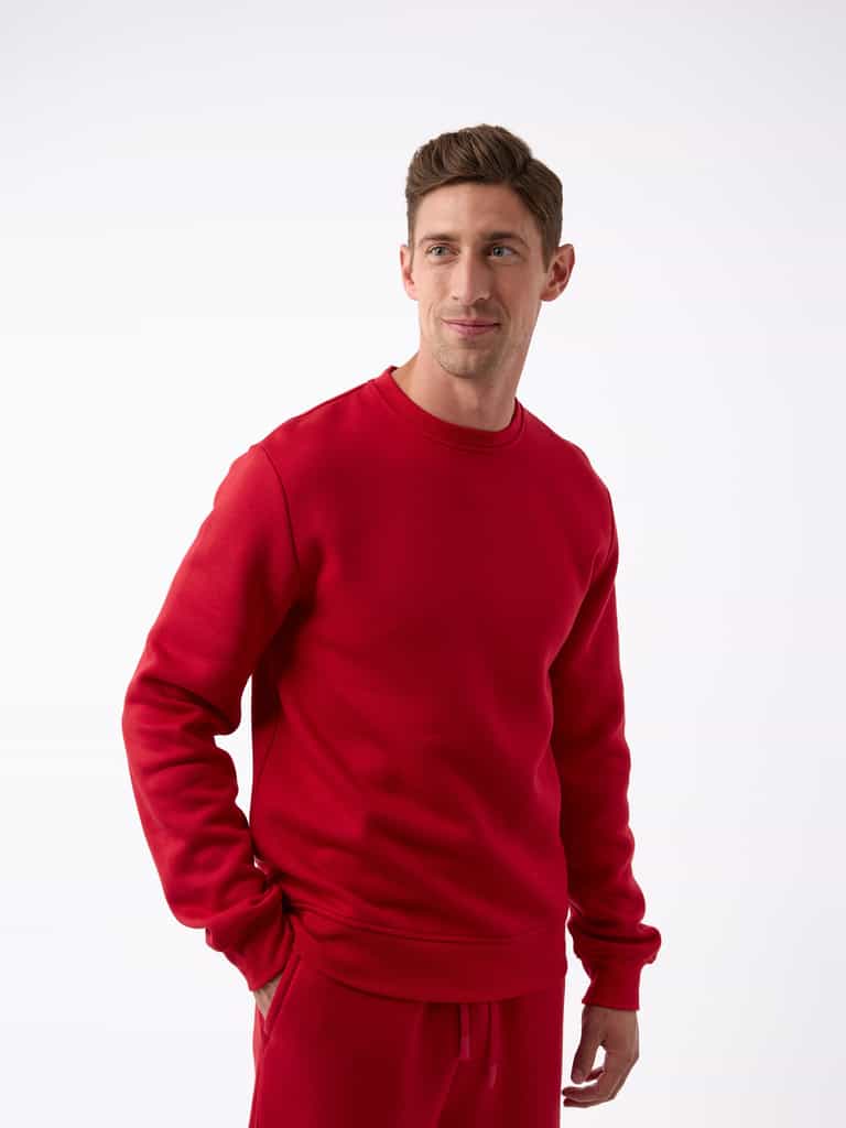 A person wearing a matching red Men's CityScape Crewneck and pants from Cozy Earth stands against a plain white background. They have short hair, are looking to the side with a slight smile, and have one hand in their pocket. |Color:Crimson