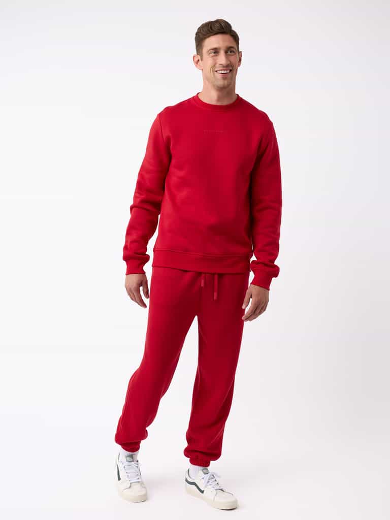 A person dressed in a matching red sweatshirt and Cozy Earth's Men's CityScape Sweatpant poses against a plain white background. They complement their outfit with white sneakers accented in green, smiling as they gaze slightly to the side. |Color:Crimson