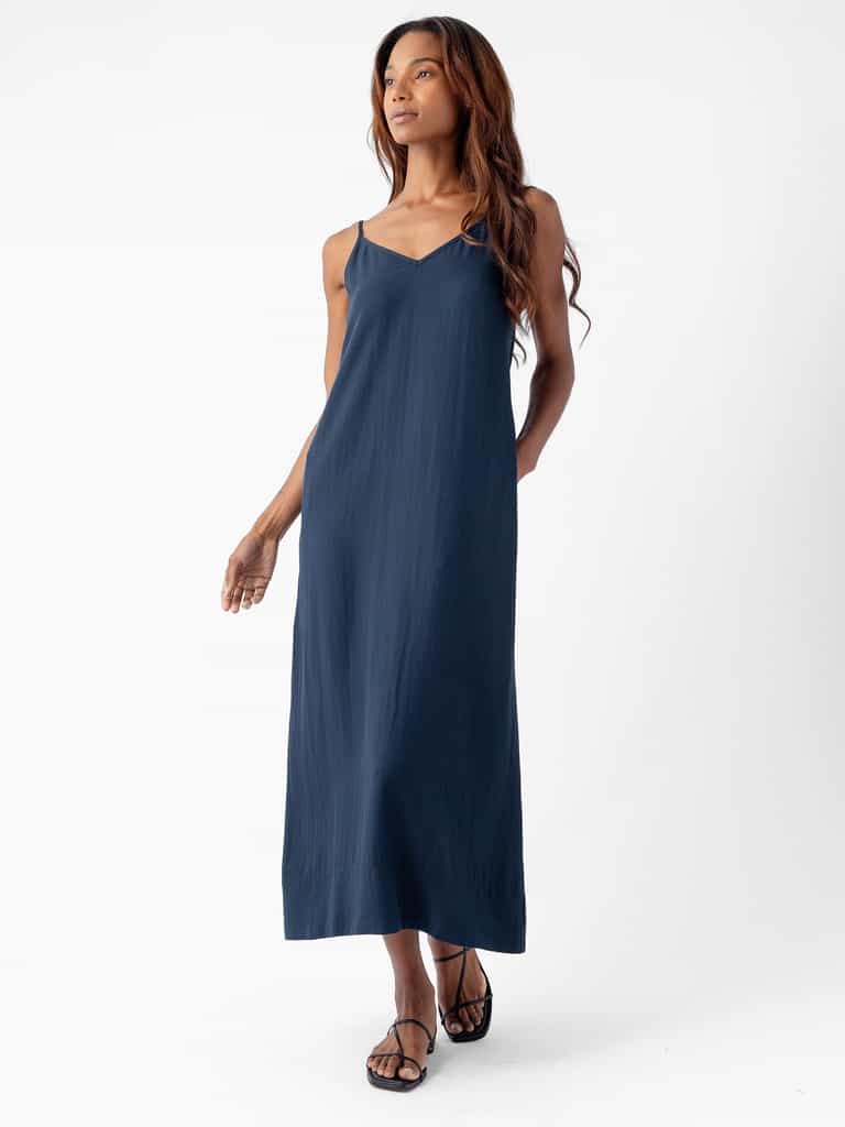 A person with long, wavy hair is wearing a Cozy Earth Women's Sunset Slip Dress in navy blue and black sandals. They are standing against a white background, looking confidently to the side with one hand in their pocket and the other arm relaxed by their side. |Color:Eclipse