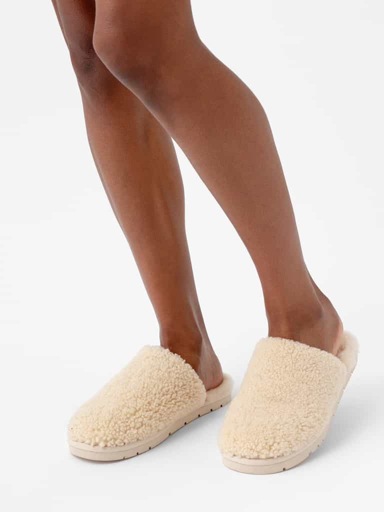 A pair of legs, shown from just below the knees, wearing Cozy Earth's Puffy Sheep Slippers in a cream-colored, fluffy slip-on style with a textured finish. The background is plain white. |Color:Natural