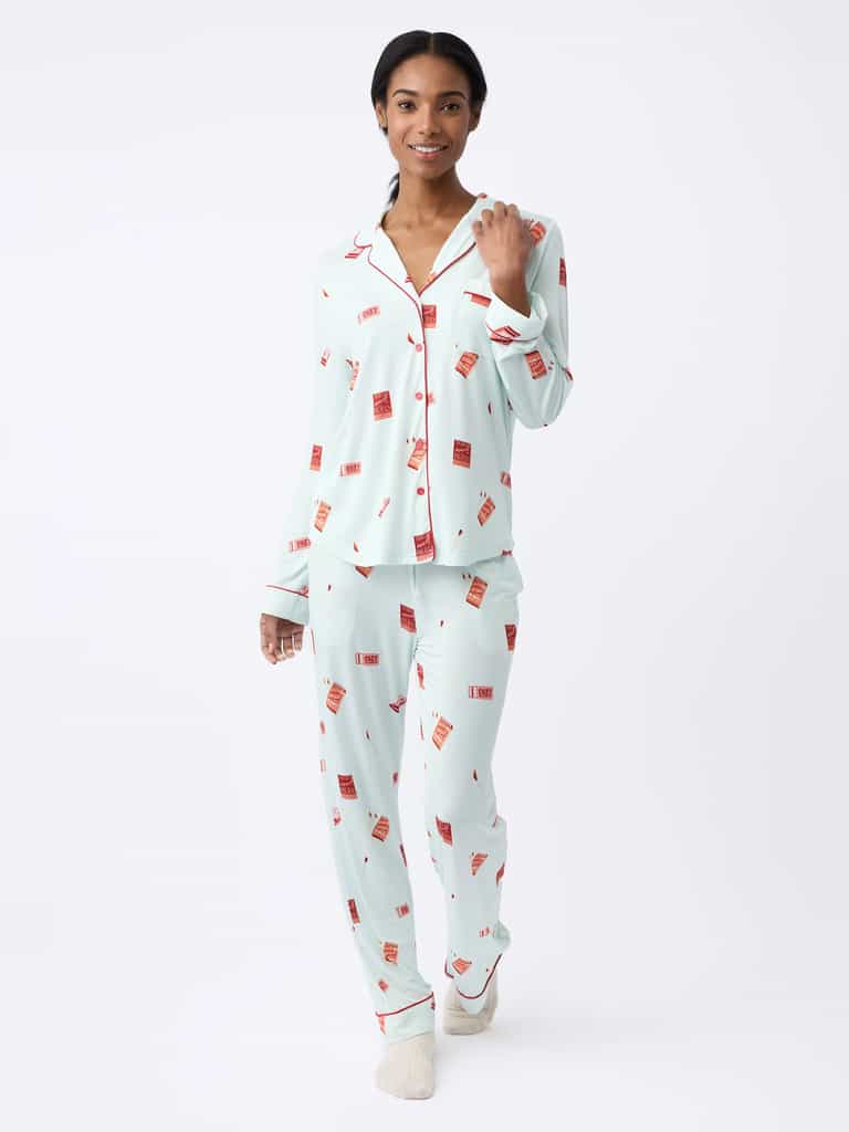 A person is joyfully walking forward, wearing the Cozy Earth Last Chance Women's Bamboo Stretch-Knit Pajama Set in light blue with red patterns and text, while holding the collar of the long sleeve top against a plain white background. |Color:Perfect Match