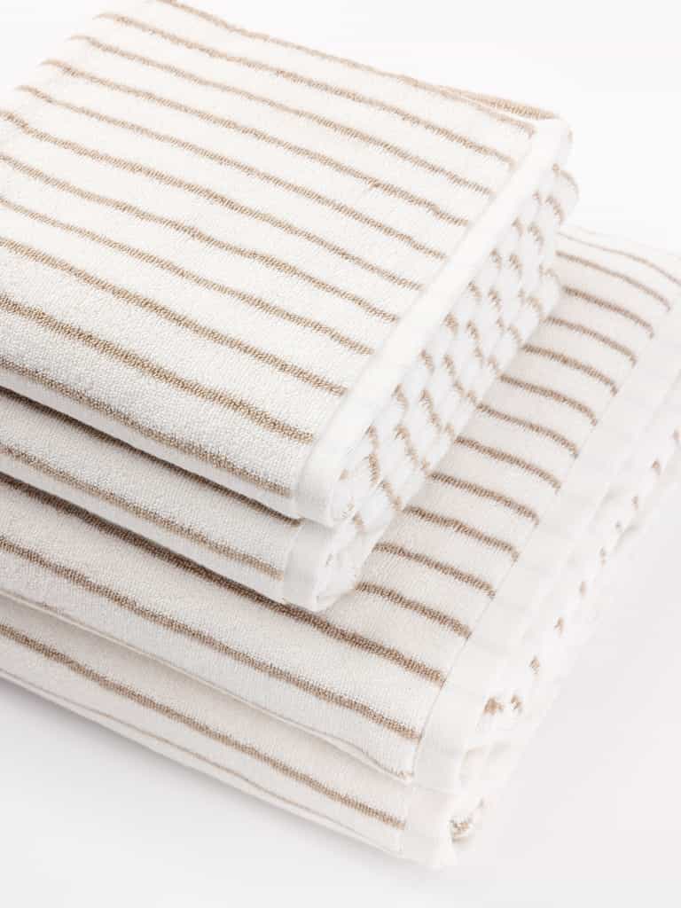 A stack of three Luxe Bath Towels by Cozy Earth in white with light brown stripes are neatly arranged, with each towel slightly offset from the one beneath it. The simple, elegant design gives a clean and inviting appearance. |Color:Sand Stripe