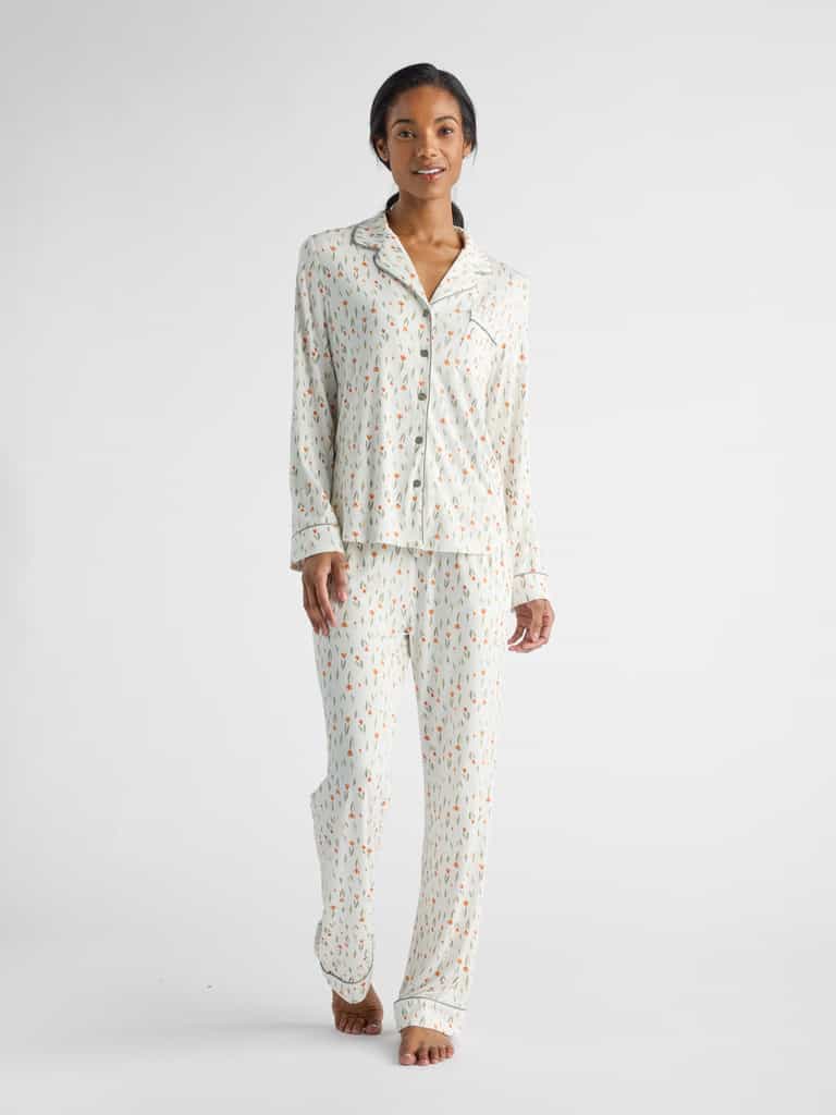 Wearing HIDE's Women's Bamboo Stretch-Knit Long Sleeve Pajama Top, a person stands barefoot on a white surface, clad in matching pajamas with a light background and small colorful patterns, smiling as they look forward.|Color:Sienna Tulips