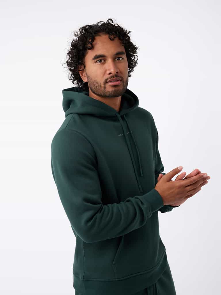 A person with curly hair is wearing a Cozy Earth Men's CityScape Hoodie in dark green and standing against a plain white background, with their hands together. |Color:Spruce