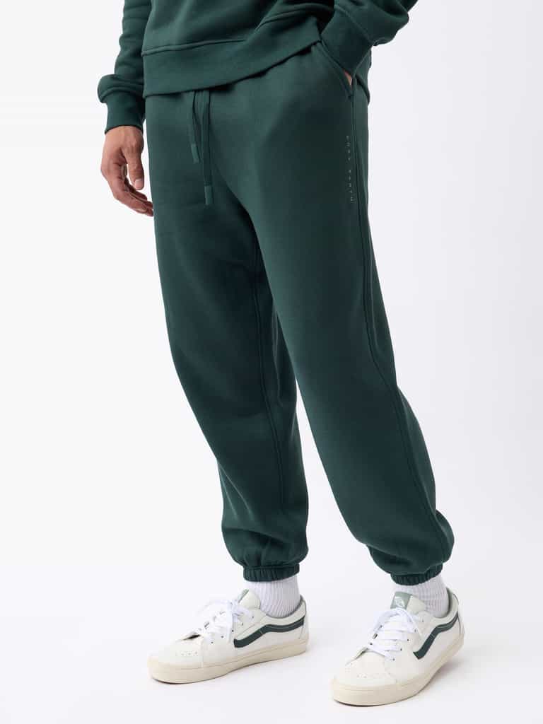 A person wearing Cozy Earth's Men's CityScape Sweatpant in dark green, paired with a matching sweatshirt. They are also sporting white sneakers featuring dark accents and white socks. The individual strikes a casual pose with their hands in pockets, set against a plain white background. |Color:Spruce