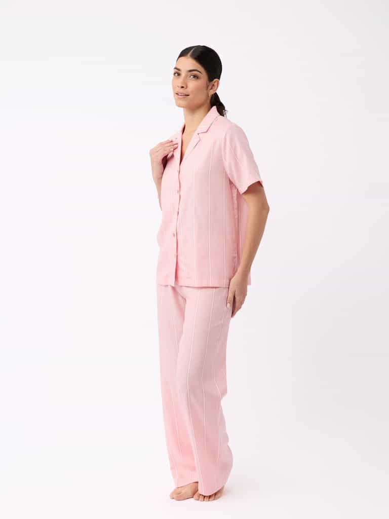 A person with long dark hair tied back smiles gently while standing barefoot against a white background, wearing Cozy Earth's Women's Soft Woven Pajama Pant set in pink with white pinstripes, featuring a matching short-sleeve button-up shirt and long pants. |Color:Sweetheart Stripe