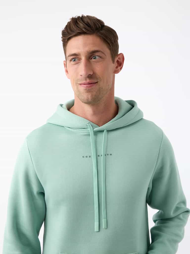 A person wearing the Men's CityScape Hoodie by Cozy Earth stands against a plain white background. The light green hoodie features drawstrings and subtle text on the chest. The person, with short brown hair, is looking slightly to the side while smiling gently. |Color:Wintergreen
