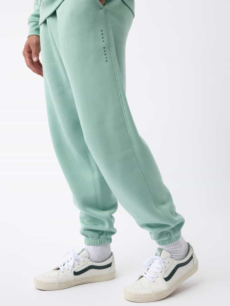 A person wearing Cozy Earth's Men's CityScape Sweatpant in pastel green, featuring subtle text detail on the side, stands casually. They complement the outfit with white sneakers accented in green and white socks against a plain white background. |Color:Wintergreen