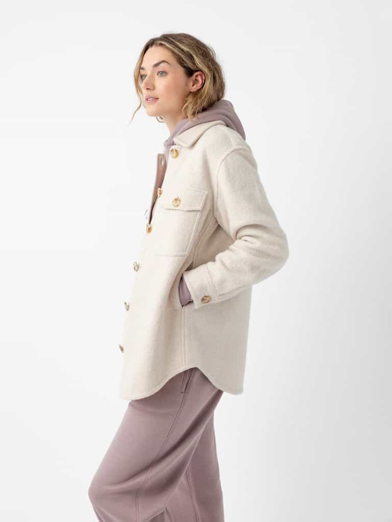 A person with shoulder-length light hair stands against a plain white background. They are wearing Cozy Earth’s Women’s Boucle Shacket in cream over a mauve hoodie, paired with matching mauve pants. The person looks to the side with a relaxed expression. |Color:Alabaster