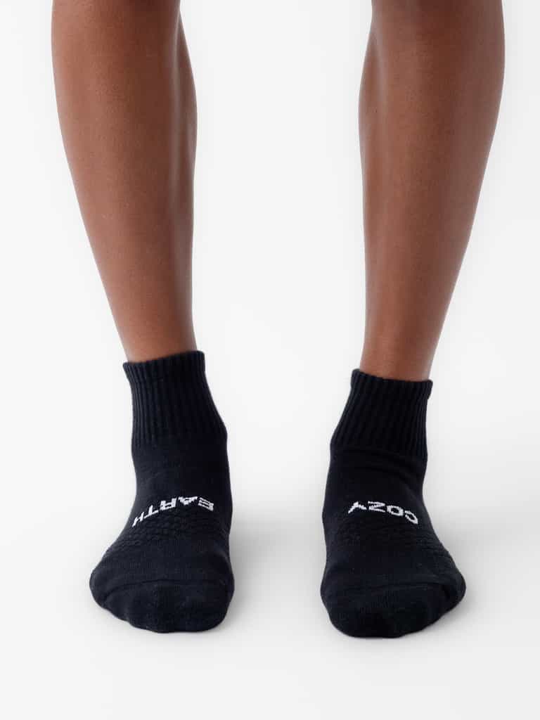 A person wears Cozy Earth Essential Quarter Socks from the 8-Pack, featuring black fabric with white text on the toes and ribbed cuffs, standing on a white background with visible lower legs. |Color:Black