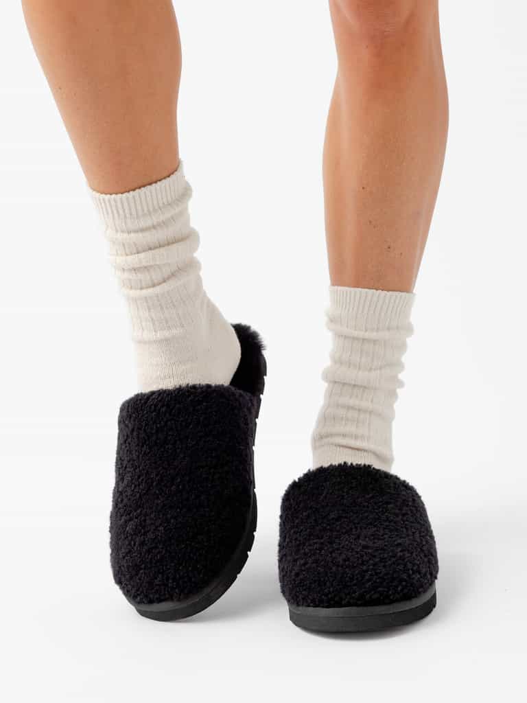 A person wearing white, ribbed socks and Cozy Earth's Puffy Sheep Slippers is standing on a white background. Only their lower legs and feet are visible. The slippers have a cozy, textured appearance that contrasts with the light-colored socks. |Color:Black Sheep