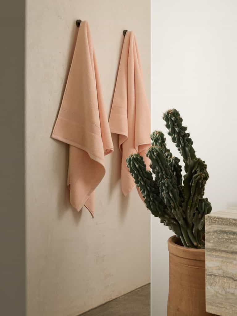 Two Luxe Bath Towels by Cozy Earth, in pink, hang side by side on a minimalist bathroom wall. Nearby is a tall cactus in a terracotta pot beside the edge of a textured stone countertop, creating a calm and stylish setting. |Color:Blossom