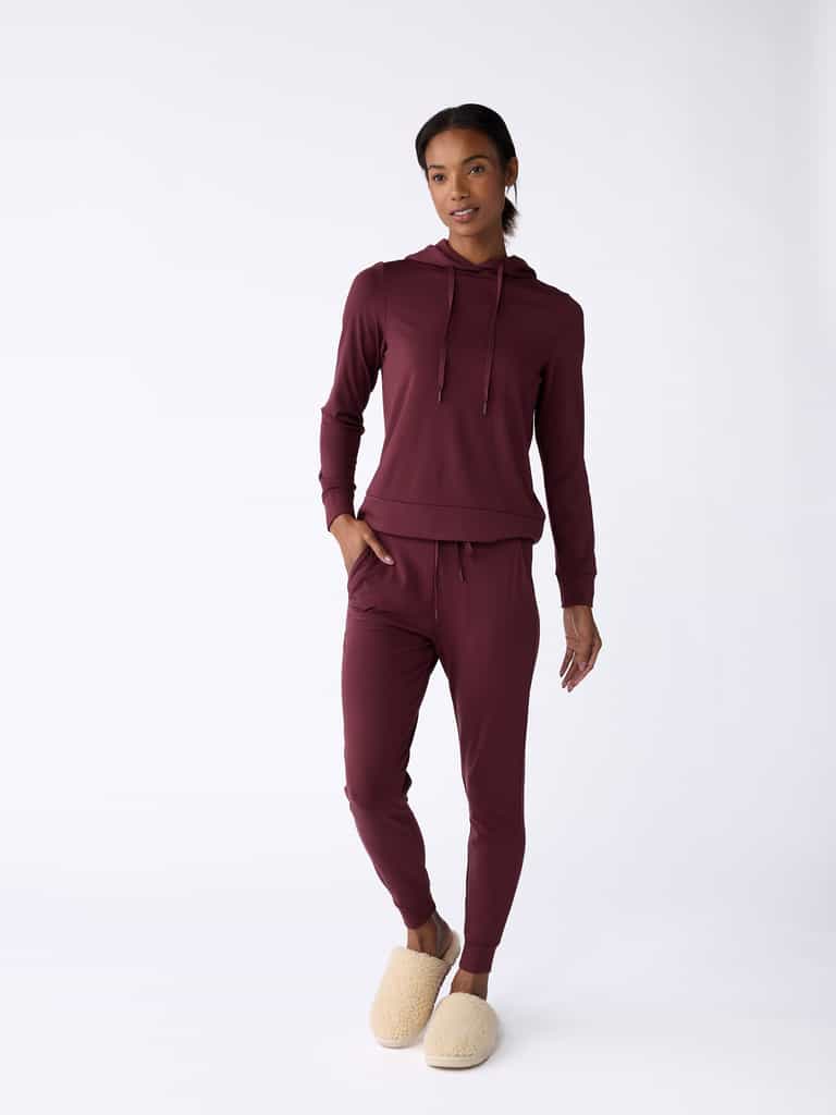 A person wearing Cozy Earth's maroon Women’s Bamboo Jogger Pant and a matching hoodie stands against a plain white background. They have their hands in their pockets and are wearing beige slippers. |Color:Burgundy