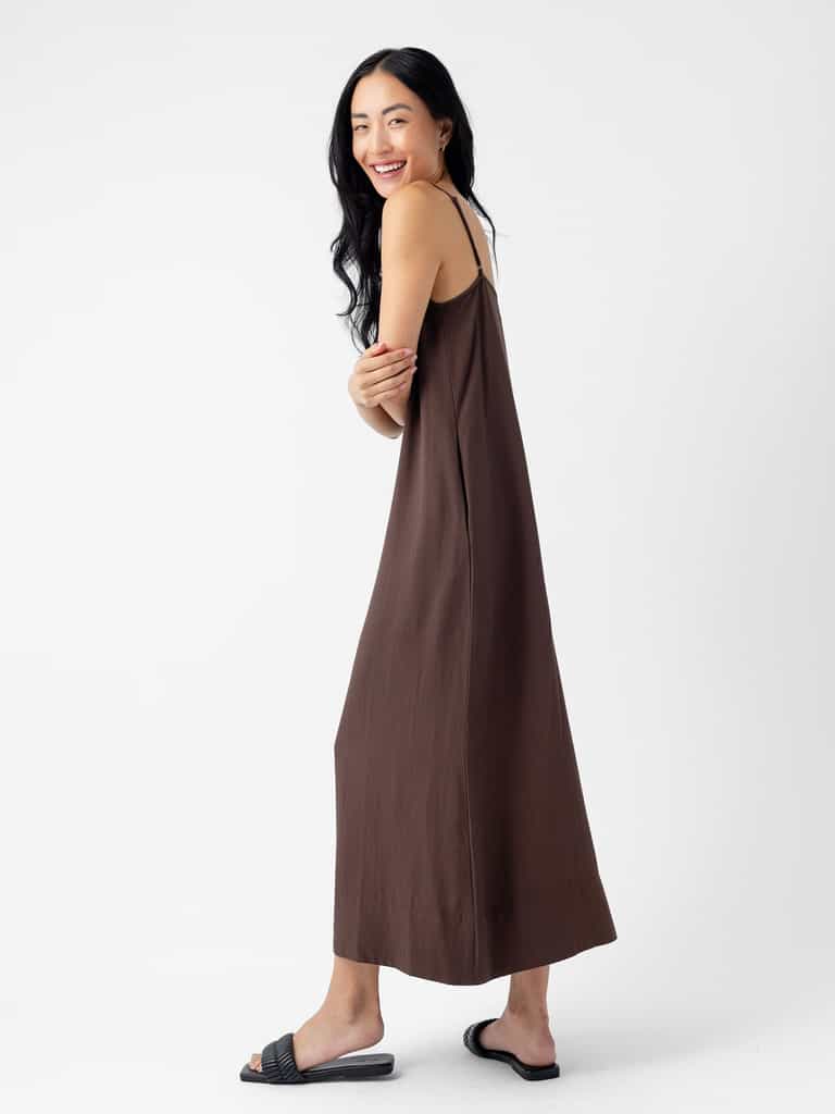 A person with long black hair smiles while wearing the Women's Sunset Slip Dress by Cozy Earth in a loose-fitting brown, sleeveless design and black slide sandals. They are standing sideways, looking over their shoulder against a plain white background. |Color:Cacao