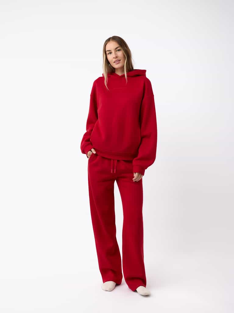 A person stands against a white background wearing a red hoodie with matching Cozy Earth's Women's CityScape Wide Leg Pant in red. They have long hair and are looking at the camera with a relaxed pose while wearing white shoes. |Color:Crimson