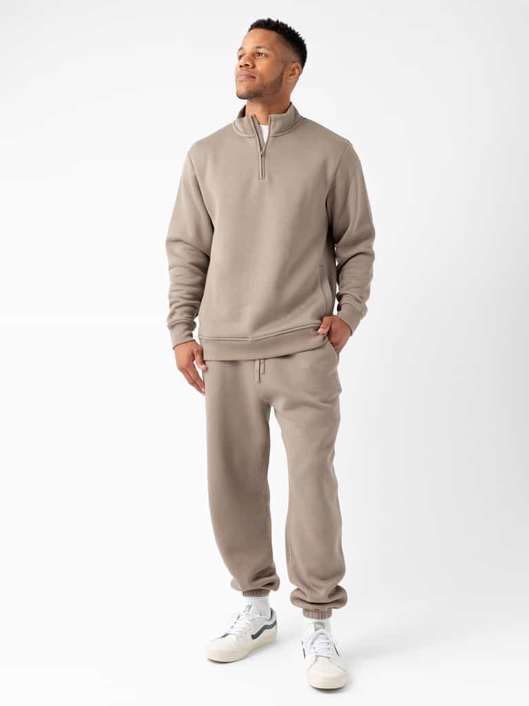 A person stands against a white background, wearing Cozy Earth's Men's CityScape Quarter Zip in taupe-colored fabric and matching sweatpants with pockets. They have their left hand in their pocket and are looking slightly upward, smiling. The outfit is paired with white sneakers. |Color:Ebony