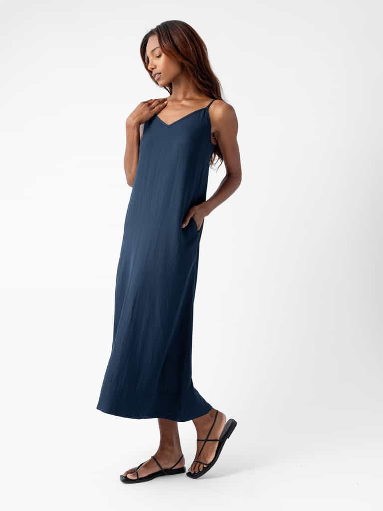 A woman with long hair is wearing the Women's Sunset Slip Dress by Cozy Earth, which features a navy blue, sleeveless, ankle-length design with a V-neckline. She is posing with one hand touching her neck and the other hand in the dress pocket while also wearing black sandals. The background is plain white. |Color:Eclipse
