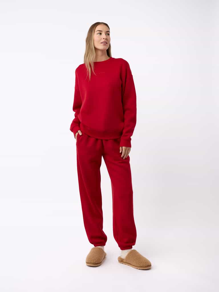 A person stands against a plain background, wearing a matching red sweatshirt and sweatpants set. They have light brown hair and are gazing to the side. On their feet, they are wearing Cozy Earth's Puffy Sheep Slippers in tan, which look fuzzy and comfortable. They have one hand in their pocket. |Color:Fawn