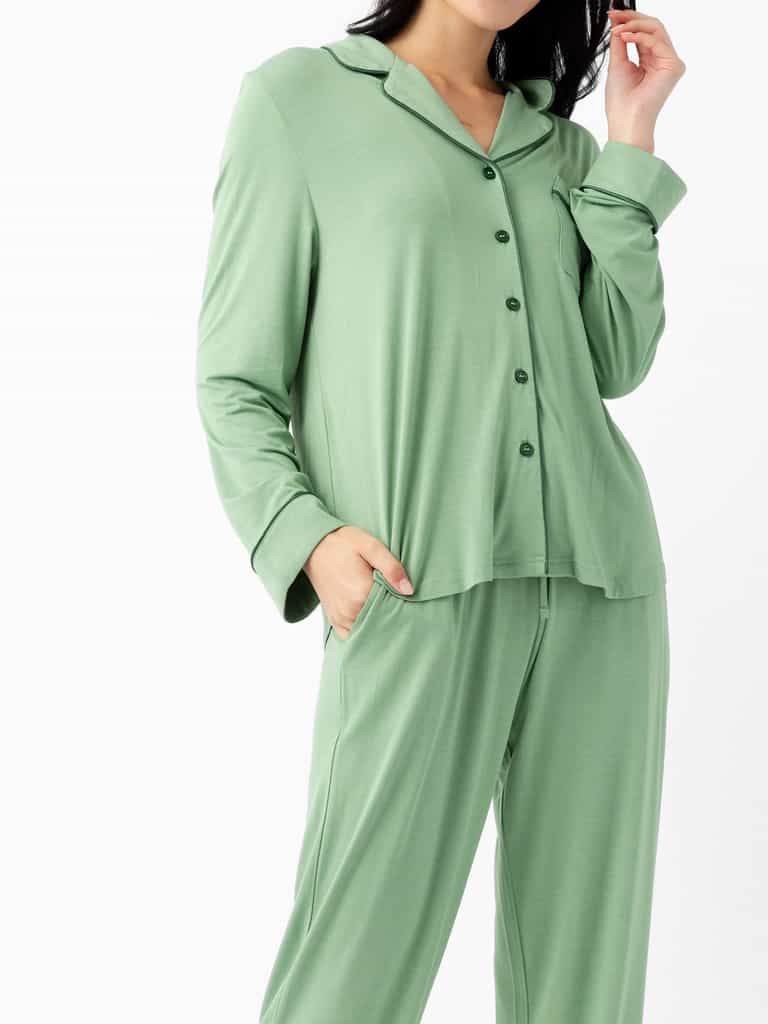 A woman wears the Cozy Earth Last Chance Women's Bamboo Stretch-Knit Long Sleeve Pajama Set in light green, featuring a loose fit, button-up top, and matching pants, with one hand in her pocket against a plain white background.