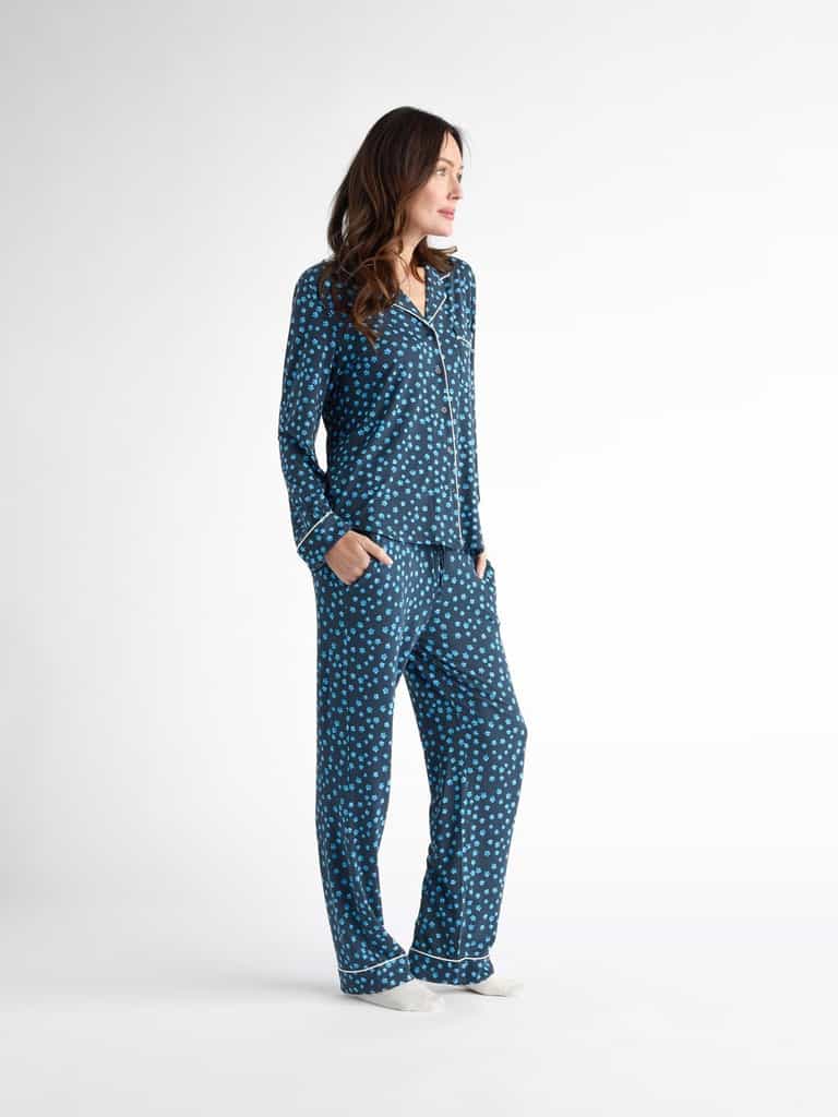 On a plain background, a person models Cozy Earth Women's Bamboo Stretch-Knit Pajama Pant in blue with white polka dots, hands in pockets, gazing to the side, with relaxed long hair and white socks. |Color:Forget Me Nots