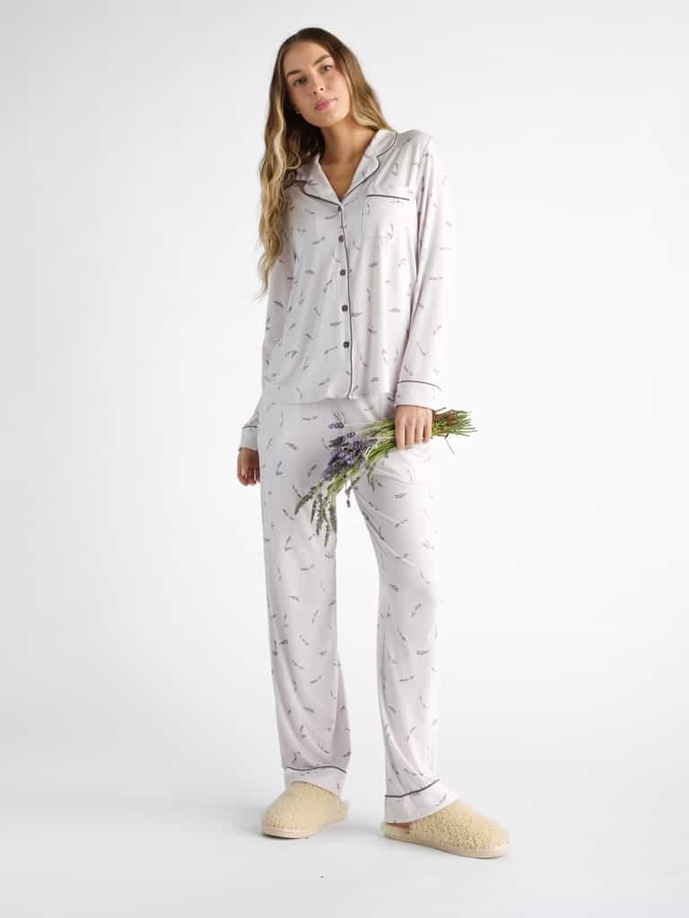 Against a plain background, a person wears Cozy earth Women's Bamboo Stretch-Knit Pajama Pant in a light color with a subtle pattern, paired with beige slippers. They hold a small lavender bouquet as their long, wavy hair cascades down. |Color:French Lavender