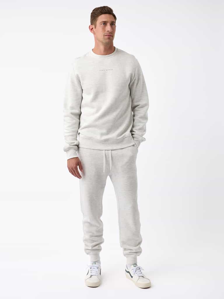 A man stands against a white background wearing a light gray Men's CityScape Jogger by Cozy Earth, paired with matching sweatpants. He is also wearing white sneakers with green accents. His hands are by his sides, and he looks slightly to his right. |Color:Heather Grey