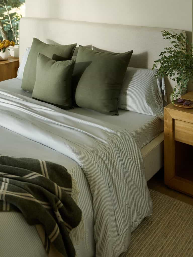 A neatly made bed boasts a light gray headboard and striped bedding, complemented by three green throw pillows. A plaid blanket is elegantly draped at the foot of the bed. Wooden nightstands are positioned on each side, while a plant introduces a touch of greenery to the scene. Enhance your sleep experience with Cozy Earth's Bamboo Flat Sheet for added comfort and style. |Color:Jade Pinstripe