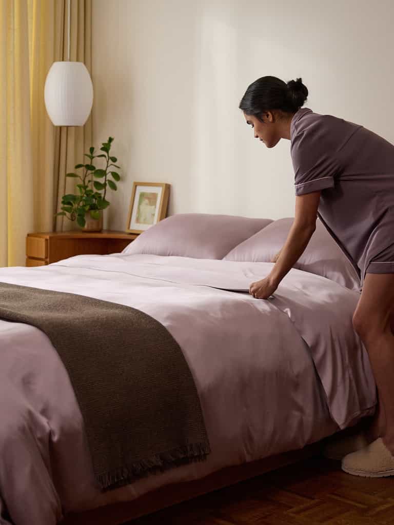 A person in pajamas makes a bed with Cozy Earth's Bamboo Flat Sheet in lavender and a brown throw blanket. Beside the bed is a wooden nightstand holding a potted plant and framed picture, while a white lamp hangs from the ceiling. |Color:Lavender