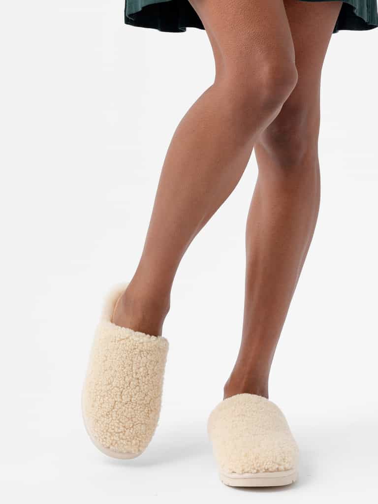 A person is wearing a dark green dress and standing with legs crossed at the ankles. Their feet are adorned in Cozy Earth's beige and fluffy Puffy Sheep Slippers, set against a white background. |Color:Natural