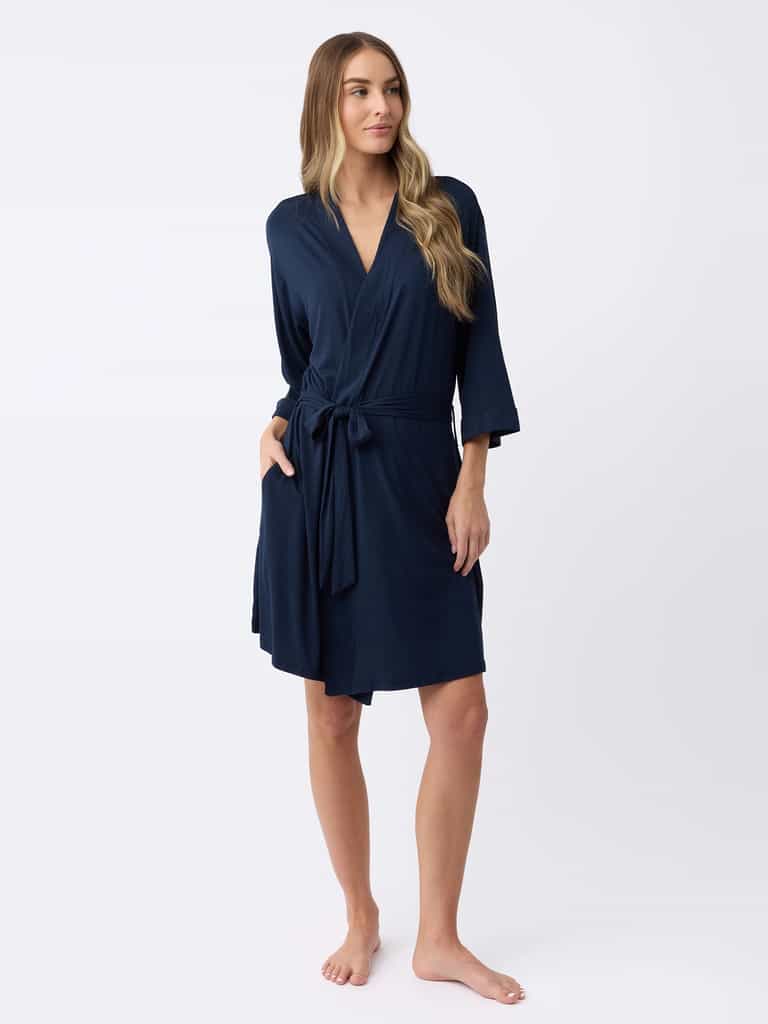A person with long hair poses barefoot against a plain white background, wearing Cozy Earth's Women's Stretch-Knit Bamboo Kimono Robe in navy blue, featuring long sleeves, a wrap style, and a waist tie. |Color:Navy