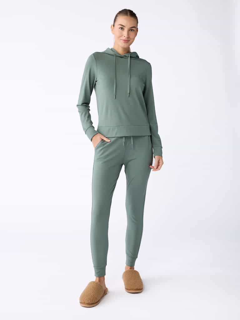 A person dressed in a light green hoodie paired with Cozy Earth's Women's Bamboo Jogger Pant stands against a plain background. They are also wearing brown fuzzy slippers, have their hair tied back, and are smiling slightly. |Color:Oakmoss